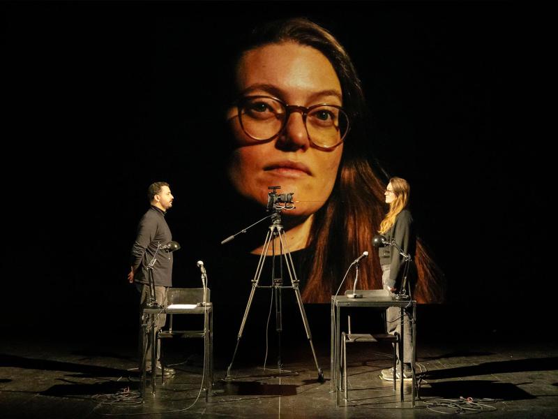 Bühnensituation. Fikri Anıl Altıntaş und Katharina Wisotzki stehen sich gegenüber, beide tragen schwarz. Neben ihnen stehen zwei schwarze Tische mit jeweils einer Lampe und einem Mikrofon drauf. Eine Kamera auf einem Stativ ist auf Katharina Wisotzki gerichtet. Das Kamerabild ihres Kopfes ist an die Wand hinter den beiden projiziert. 