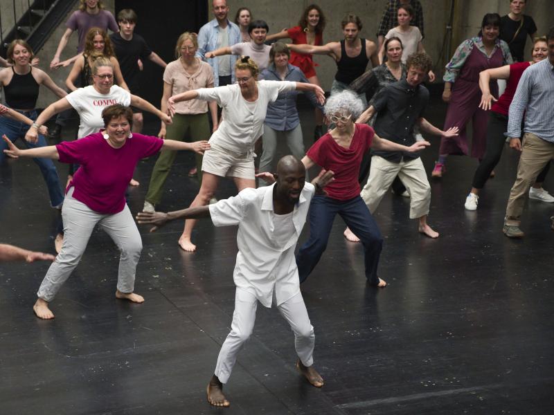 Eine Gruppe älterer Personen steht verteilt auf der Bühne des MalerSaals. Sie machen alle die gleichen oder ähnlichen Bewegungen: die Beine auseinander, die Knie gebeugt und die Arme zu beiden Seiten ausgestreckt. Viele lächeln, andere gucken konzentriert. Sie tragen sportliche, sommerliche Kleidung.