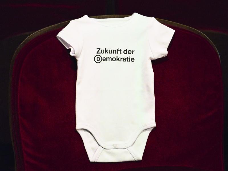 Ein weißer Baby-Body hängt über einen roten Samtsitz des Schauspielhauses. Mittig auf der Brust steht „Zukunft der Demokratie“