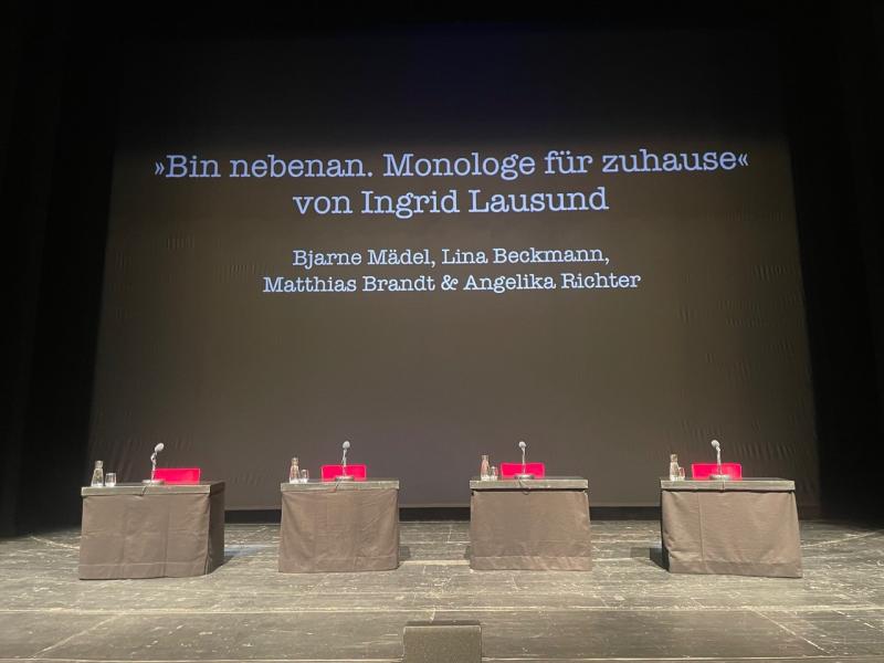 Bühnensituation. Vier Pulte mit vier roten Stühlen dahinter. Dahinter eine schwarze Leinwand mit dem Schriftzug "Bin nebenan. Monologe für zuhause"