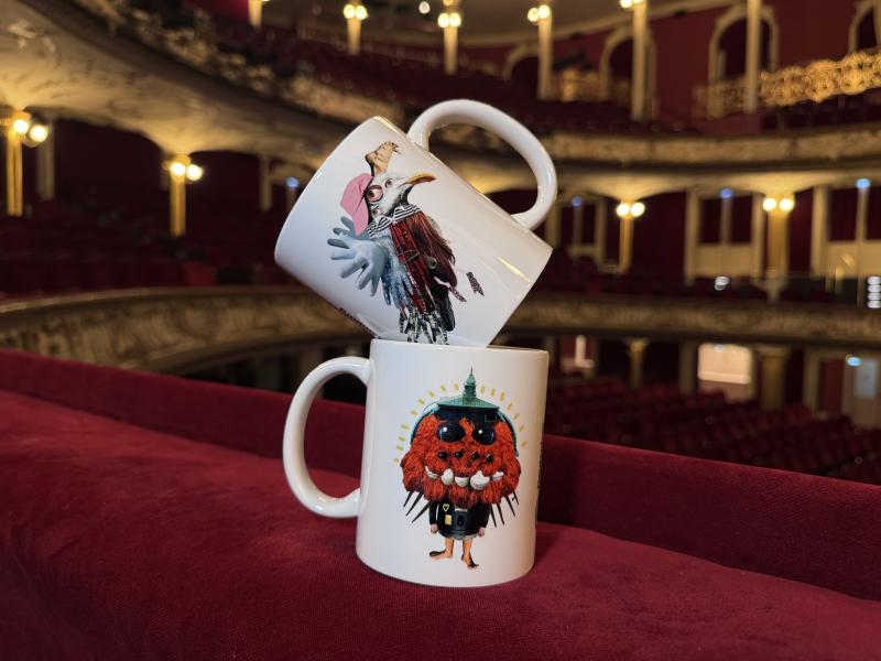 Zwei weiße Tassen mit den bunten Spielzeitmotiven des SchauSpielHauses stehen auf einer roten Unterlage aus Samt. Im Hintergrund der Saal des SchauSpielHauses.