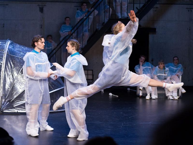 Ein junger Mann und eine junge Frau mit geflochtenen Zöpfen unterhalten sich. Im Vordergrund macht eine weitere junge Frau mit geflochtenen Zöpfen einen Spagat in der Luft. Im Hintergrund sitzen drei Personen auf einer Bank, sie beobachten die Personen im Vordergrund. Noch weiter im Hintergrund stehen weitere Personen auf einer Treppe, die das Geschehen beobachten. Alle Personen auf der Bühne des MalerSaals tragen durchsichtige Maleranzüge.