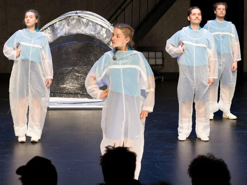 Auf der Bühne des MalerSaals stehen vier Personen in durchsichtigen Maleranzügen versetzt nebeneinander. Sie haben alle ihre rechte Hand in die Hüfte gestemmt. Im Hintergrund steht ein durchsichtiges Iglu-Zelt aus Plastik.