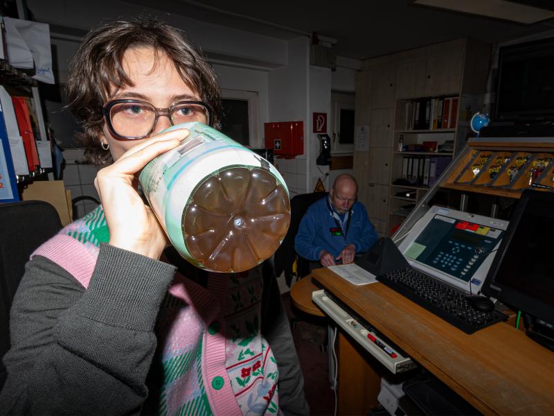 Eine Person steht dicht an der Kamera und hält eine große Plastikflasche zum Trinken an den Mund. Im Hintergrund ist der Arbeitsbereich der Bühnenpforte eines Theaters zu sehen. An einem Tisch sitzt eine weitere Person in einem blauen Oberteil. Um sie herum Regale, Mappen, Ordner, Briefumschläge, ein Pult und ein Schlüsselbrett.