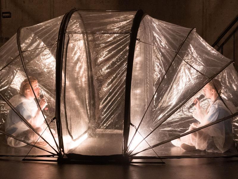 Auf der Bühne des MalerSaals steht ein durchsichtiges Iglu-Zelt aus Plastik. Es ist von innen beleuchtet. Darin sitzen sich zwei Personen gegenüber im Schneidersitz, beide halten sich ein Mikrofon an den Mund. Sie tragen beide durchsichtige Maleranzüge.