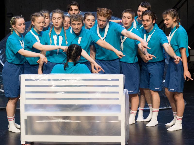 Auf einer weißen Bank, die in Plastik eingehüllt ist, sitzt eine junge Frau im türkisen T-Shirt mit dem Rücken zum Publikum. Vor ihr stehen dreizehn Personen, alle tragen türkise T-Shirts. Sie zeigen alle mit dem Finger und ausgestreckten Armen auf den Boden rechts neben der Bank. Dabei schauen sie alle die junge Frau auf der Bank an, manche haben den Mund geöffnet.