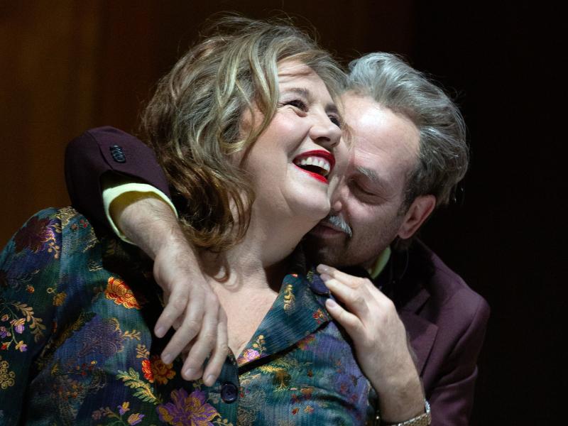 Bühnensituation. Bettina Stucky und Samuel Weiss sitzen auf einem grünen Sofa. Bettina trägt lockiges, schulterlanges Haar, roten Lippenstift, lacht und schaut an der Kamera vorbei. Sie trägt einen dunkelgrünen Samtanzug mit bunten Blumen-Applikationen drauf. Neben ihr sitzt Samuel, der einen Arm um ihre Schulter und die andere Hand auf ihre andere Schulter gelegt hat. Er dreht sein Gesicht leicht in ihre Haare, lächelt und hat die Augen geschlossen.