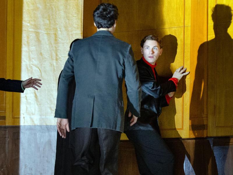 Bühnensituation. Paul Behren steht in einem großen, holzvertäfelten Raum und trägt einen schwarzen Anzug mit roten Akzenten. Sein Körper ist zur Wand gewannt, doch er schaut ins Publikum. Pascal Houdus geht auf ihn zu, mit dem Rücken zum Publikum. Er trägt einen schwarzen Anzug.