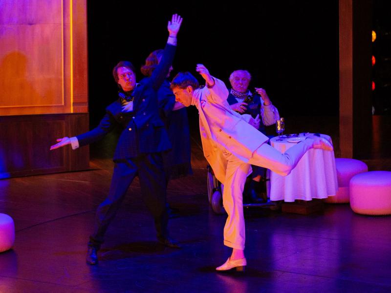 Paul Behren trägt einen weißen und Daniel Hoevels einen schwarzen Anzug. Sie bewegen sich in einem holzgetäfelten Raum mit erhobenen Armen und tanzen. Das Licht wirft bunte Farben auf die Bühne. Josef Ostendorf und Bettina Stucky sind in Hintergrund zu sehen. Josef sitzt in einem Rollstuhl und raucht. Neben ihm steht ein gedeckter Tisch mit weißer Tischdecke.