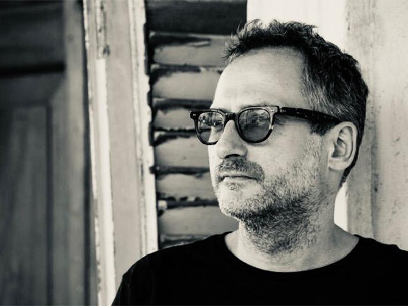 Schwarz-Weiß-Foto. Der Autor Roland Schimmelpfennig steht vor einer Hauswand. Er schaut an der Kamera vorbei. Er trägt ein dunkles Oberteil, kurze Haare und eine dunkle Sonnenbrille.