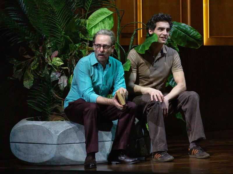 Samuel Weiss und Pascal Houdus sitzen in einem großen, holzvertäfelten Raum auf dem Rand eines großen Blumentopfes, aus dem palmenartigen Blätter herausragen. Eins der Blätter berührt Pascal Houdus Schulter. Samuel trägt ein türkises Polohemd und eine braune Hose und Pascal ein braunes Polohemd und eine braune Hose. Samuel hält ein Buch in den Händen und blickt zur Seite.