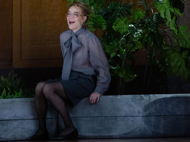 Angelika Richter sitzt in einem großen, holzvertäfelten Raum auf dem Rand eines großen Blumentopfes, aus dem palmenartigen Blätter herausragen. Sie trägt eine Brille, eine graue Bluse mit einer Schleife vorne dran und einen schwarzen Rock und Strumpfhose. Sie lacht mit offenem Mund.