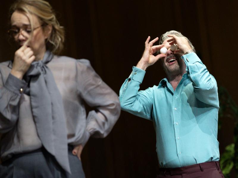 Samuel Weiss hält sich mit geöffnetem Mund zwei Tischtennisbälle vor die Augen. Er trägt ein türkises Hemd. Vor ihm steht Angelika Richter, die sich die Hand vor den Mund hält und die Stirn runzelt. Sie trägt eine Brille und eine graue Bluse mit einer Schleife vorne dran.
