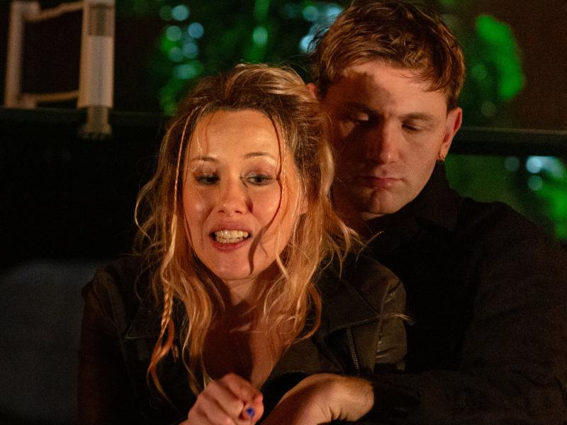 Paul Behren sitzt mit Josefine Israel auf den Boden. Er hält sie in den Armen. Paul guckt auf dem Boden. Josefine zeigt auf etwas, dabei hat sie den Mund leicht geöffnet und den Blick abgewandt. Im Hintergrund sieht man einen großer Blumentopf, aus dem palmenartige Blätter herausragen.