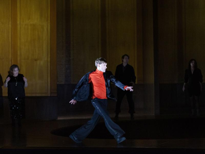Bühnensituation. Paul Behren ist im Vordergrund, er geht mit großen Schritten über die Bühne.  Er trägt einen schwarzen Anzug mit einem roten Oberteil darunter. Im Hintergrund stehen Bettina Stucky, Daniel Hoevels und Henni Jörissen, die in schwarz gekleidet sind. 