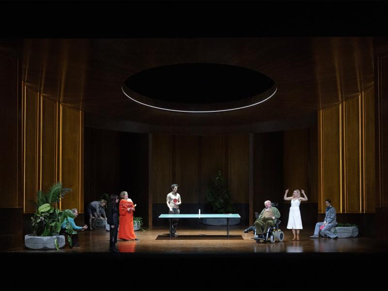 Bühnensituation. Das Ensemble in einem großen, holzvertäfelten Raum. In der Mitte eine Tischtennisplatte. Die Ensemblemitglieder stehen verteilt auf der Bühne um die Tischtennisplatte herum. Manche halten Tischtennisschläger in den Händen, andere sitzen abseits auf dem Rand großer Blumentöpfe.