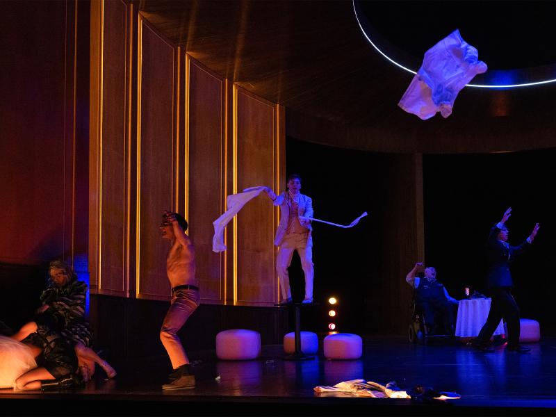 Bühnensituation. Josef Ostendorf, Pascal Houdus, Daniel Hoevels und Paul Behren sind in einem großen, holzvertäfelten Raum. Sie bewegen sich sehr ausgiebig. Paul Behren tanzt in einem weißen Anzug auf einem Tisch. Pacal Houdus tanzt ohne Oberteil. Henni Jörissen hockt auf dem Boden und ist nach vorne gebeugt, Angelika Richter hält ihren Kopf.