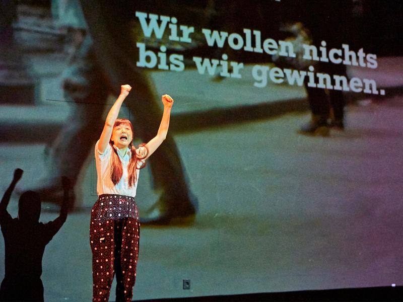 Bühnensituation. Sachiko Hara steht vor einer großen Leinwand, darauf sind Fotos projiziert. Dort steht der Satz "Wir wollen nichts bis wir gewinnen." Sachiko hat bei Hände erhoben und zur Faust geballt. Sie trägt zwei lange dunkle Zöpfe, ein weißes Shirt und eine dunkle Hose und hat den Mund geöffnet, als würde sie etwas rufen.