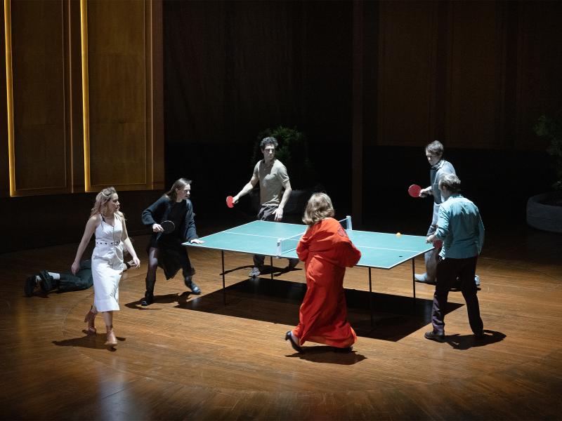 Bühnensituation. Josefine Israel, Henni Jörissen, Pascal Houdus, Daniel Hoevels, Samuel Weiss und Bettina Stucky haben alle Tischtennisschläger in den Händen und laufen um eine Tischtennisplatte herum. Im Hintergrund hohe Wände, die holzvertäfelt sind. Auch der Boden ist holzvertäfelt. Links am Rand liegt Paul Behren auf dem Boden.