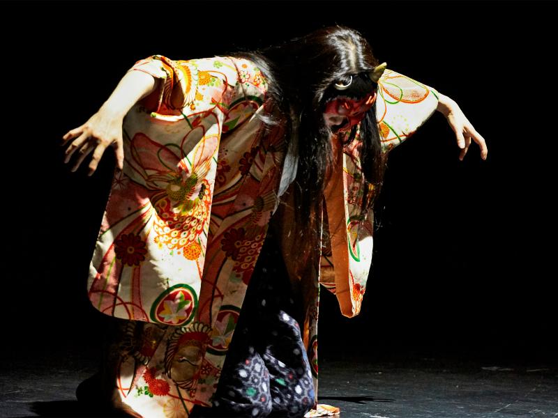 Bühnensituation. Sachiko Hara kniet auf dem Biden, sie ist nach vorne gebeugt und hebt die Arme auf Schulterhöhe, als hätte sie Flügel. Sie trägt offene, lange dunkle Haare, eine dunkle Hose und eine bunte weite Jacke. Ihr Gesicht ist zum Boden gerichtet und nicht zu erkennen.