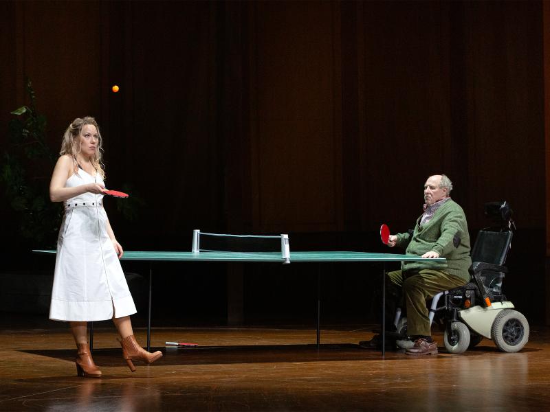 Bühnensituation. Josefine Israel steht an einer grünen Tischtennisplatte. Sie trägt braune Lederstiefel mit Absatz und ein weißes schulterfreies Kleid. Sie hält einen Tischtennisschläger in der rechten Hand und lässt einen Ball darauf springen. Ihr Blick ist auf den Ball in der Luft gerichtet. Am anderen Ende der Tischtennisplatte sitzt Josef Ostendorf in einem Rollstuhl. Er trägt eine grüne Hose, braune Halbschuhe und eine grüne Strickjacke über einem Hemd. In der rechten Hand hält er einen Tischtennisschl