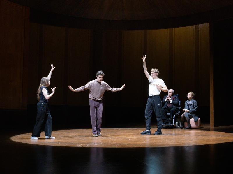 Bühnensituation. Henni Jörissen, Pascal Houdus und Paul Behren stehen in einem holzgetäfelten Raum. Sie haben die Arme erhoben und tanzen. Ein runder Kegel aus Licht ist nur auf sie gerichtet. Am Rand des Lichtkegels sitzen Angelika Richter auf einem rosafarbenen Pouf und Josef Ostendorf in einem Rollstuhl. Beide betrachten die Tanzenden.