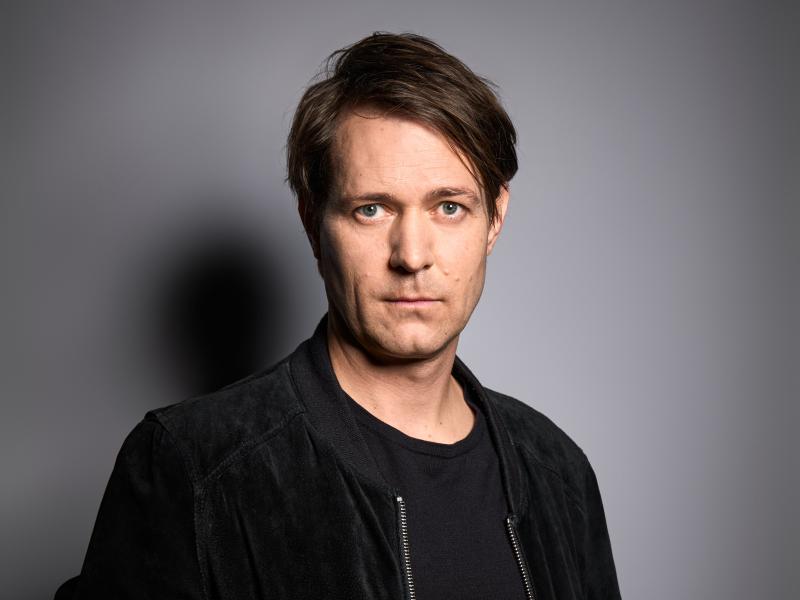 Porträt. Der Schauspieler Daniel Hoevels steht vor einem grauen Hintergrund. Er trägt eine schwarze Jacke und ein schwarzes T-Shirt. Er trägt kurze, mittelbraune Haare und schaut in die Kamera.