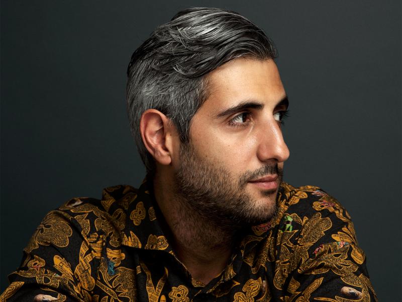 Michel Abdollahi sitzt vor einem grauen Hintergrund. Er trägt ein schwarzes Hemd mit buntem Aufdruck und kurze, mittelgraue Haare. Er schaut nach links.