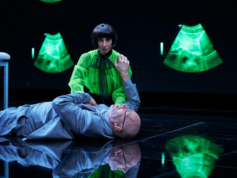Szenenfoto: Joachim Meyerhoff liegt in einem grauem Anzug auf einem glatten spiegelnden Boden. Neben ihm kniet Zeynep Buyraç mit schwarzer Bob-Frisur und einer neongrünen weiten Bluse. Ihre Hand liegt auf der Brust von Meyerhoff, dieser hält die Schulter von Buyraç fest. Der Hintergrund ist schwarz, an der Rückwand sind neongrüne trichterförmige Abbildungen zu sehen.