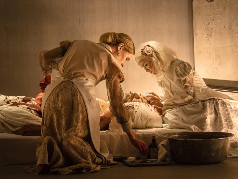 Szenenfoto: Zwei Frauen in heller, eleganter mittelalterlicher Kleidung beugen sich über einen Mann, der auf einer Matratze auf dem Boden liegt. Er ist blutbeschmiert, wie auch das Bettzeug. Die Frauen tupfen ihm die Stirn ab, seitlich stehen Medikamente und eine große Metallschüssel.
