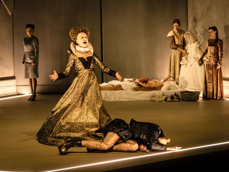 Szenenfoto: Sieben Personen auf einer Bühne. Im Vordergrund liegt eine Person am Boden, hinter ihr eine weitere Person mit einem weitem goldenen Kleid und ausgebreiteten Armen, ihr Mund ist geöffnet. Im Hintergrund ein Mann auf einer blutverschmierten Matratze, daneben drei Frauen in hellen und braunen Kleidern. Eine weitere Person steht etwas Abseits und schaut nach vorn.