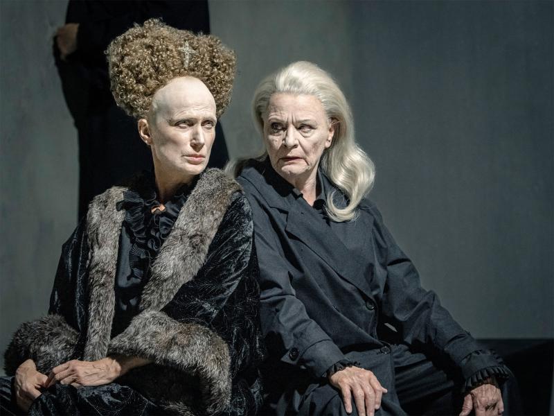 Szenenfoto: Zwei Personen lehnen sich mit den Schultern aneinander. Die linke Person trägt einen schwarzen Samtumhang mit Fellkragen, ihr Gesicht ist weiß geschminkt, sie trägt eine hochgesteckte Lockenfrisur mit sehr hoher Stirn. Die rechte Person hat lange offene graue Haare und trägt schwarze Kleidung. Sie schaut zu der linken Person, ihr Mund ist geöffnet.