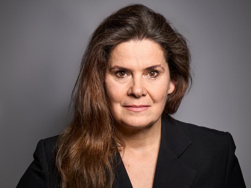 Julia Wieninger steht vor einem grauen Hintergrund. Sie trägt lange, dunkelbraue Haare und einen schwarzen Blazer. Sie schaut in die Kamera.