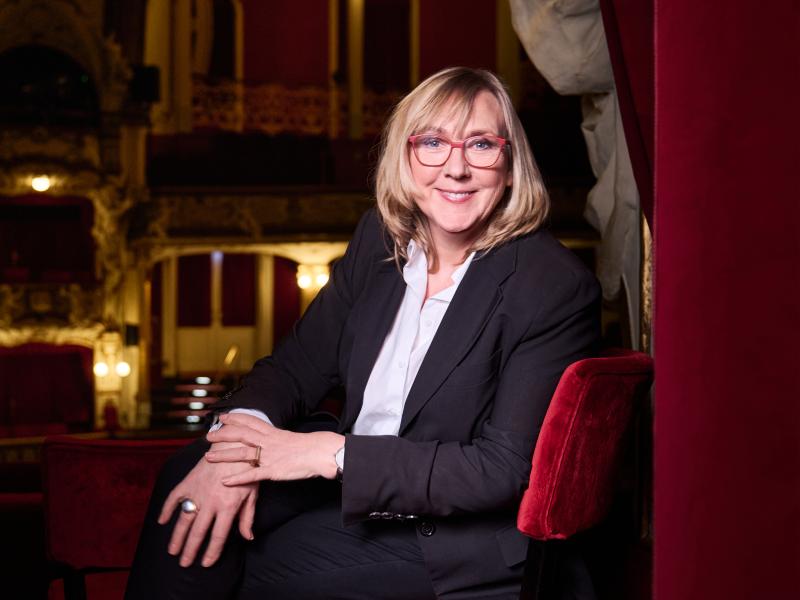 Kathrin Hartkopf sitzt in einer Loge im Saal des Schauspielhauses auf einem roten Samtstuhl. Sie trägt schulterlange blonde Haare, eine rote Brille, eine helle Bluse, einen dunklen Blazer und eine dunkle Hose. Im Hintergrund ist der Saal des Theater mit seinen roten Sitzen zu sehen. 