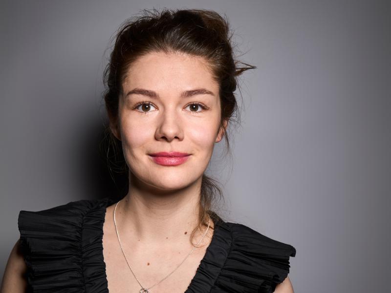 Linn Reusse steht vor einem grauen Hintergrund. Sie trägt ein schwarzes Oberteil, eine filigrane Kette und pinken Lippenstift. Sie hat die Haare zurückgebunden und lächelt in die Kamera.