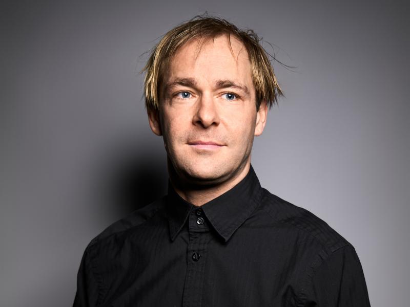 Mirco Kreibich steht vor einem grauen Hintergrund. Er trägt ein schwarzes, bis oben zugeknöpftes Hemd und kurze blonde Haare. Er schaut in Richtung der Kamera aber leicht daran vorbei.