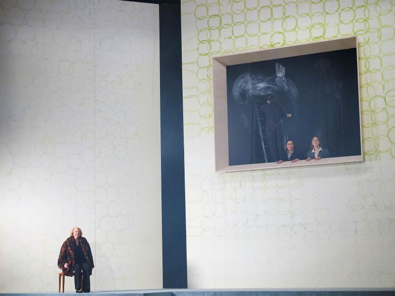 Bühnensituation: Ilse Ritter sitzt auf einem Stuhl. Im Hintergrund zwei hohe weiße Wände mit aufgemalten grünen Kreisen. Eine Wand hat eine große rechteckige Öffnung. Aus der Öffnung gucken Alberta von Polnitz und Julia Wieninger heraus. Hinter ihnen ein Mann im schwarzen Anzug auf einer Leiter, dahinter verwischte Kreidespuren an der Wand.