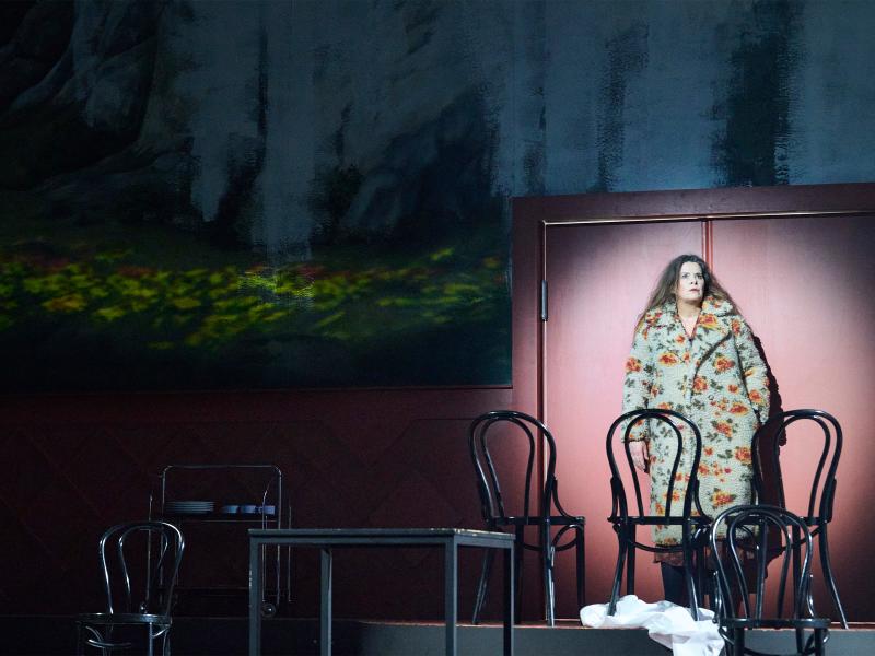 Julia Wieninger steht mit einem geblümten Filzmantel vor einer roten Tür. Vor ihr stehen drei schwarze Stühle mit Lehne, ein Tisch. Im Hintergrund ein Servierwagen mit Tellern und Tassen. 