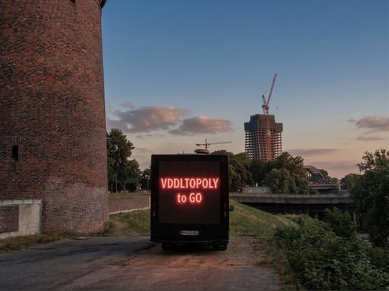 Außenansicht. Links im Bild ist ein großes rotes Backsteingebäude zu erkennen. Davor, auf einem rauen Betonuntergrund, parkt ein schwarzer LKW mit einer großen Leuchtschrift auf der Rückseite. Dort steht: VDDLTOPOLY to GO. Im Hintergrund ist eine Flussumgebung des Hamburger Hafens mit einer Brücke und Autos zu sehen. Ganz im Hintergrund ragt der unfertige bau des Elbtowers mit zwei großen Kränen in den Himmel.