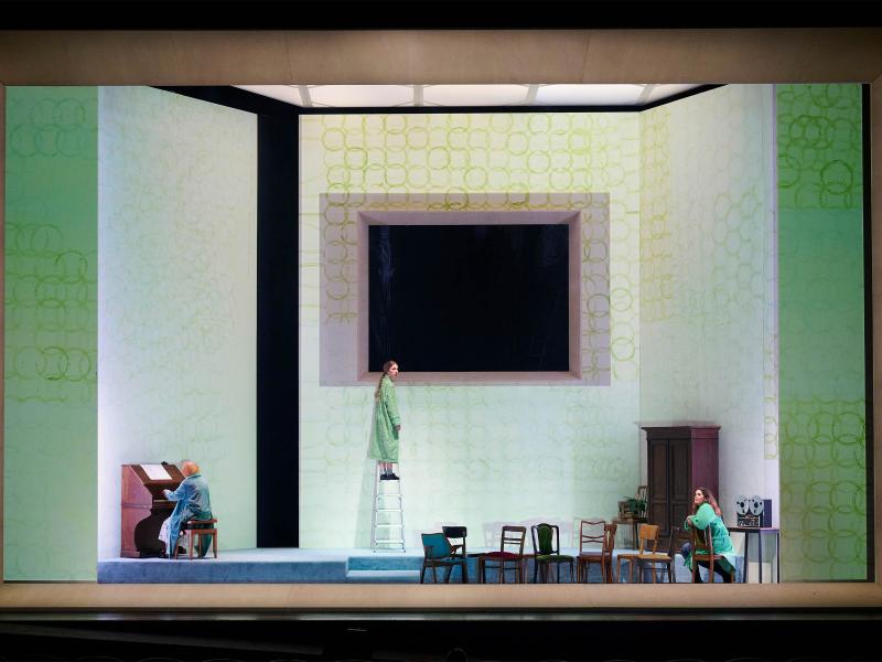 Bühnensituation: Das Bühnenbild besteht aus hohen weißen Wänden auf denen grüne überlappende Kreise aufgemalt sind. Die mittlere Wand hat eine große rechteckige Öffnung, dahinter ist es schwarz. Ilse Ritter sitzt mit dem Rücken zum Publikum links an einem Klavier. Mittig steht Alberta von Polnitz auf einer Leiter. Rechts sitzt Julia Wieninger auf einem Stuhl. Alle drei tragen grün-blaue Bademäntel. Mehrere Stühle sind auf der Bühne verteilt. Rechts ein Holzschrank sowie ein Tonbandgerät auf einem Tisch.