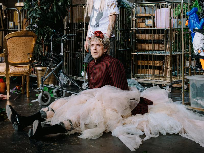 Bühnensituation. Christoph Jöde sitzt auf dem Boden und trägt einen großen Tüllrock und eine rot-schwarz gestreifte Jacke und schwarze Stiefel. Hinter ihn stehen zahlreiche Wagen mit Requisiten drauf. Links hinter ihm steht ein goldener Stuhl. Direkt hinter ihm steht eine weitere Person. Ihr Gesicht ist nicht zu sehen, man sieht nur ihr Arme, ihre Beine und einen Teil ihres Oberkörpers. Sie trägt ein weißes T-Shirt und eine gestreifte kurze Hose.