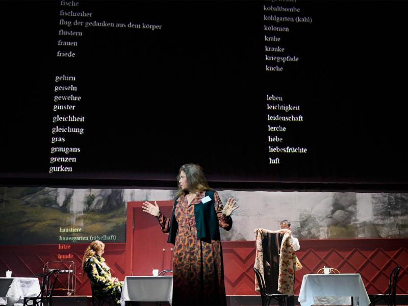 Bühnensituation: Julia Wieninger steht im Vordergrund und schaut zur Seite, die Hände erhoben. Hinter ihr drei Tische mit weißen Tischdecken und weißen Bechern darauf. Alberta von Polnitz sitzt an dem mittleren. Über ihnen eine schwarze Leinwand, auf die weißer Worte untereinander projiziert werden: fische, fischreiher, flug der gedanken aus dem körper, flüstern, frauen, friede, gehirn, geiseln, gewehre …