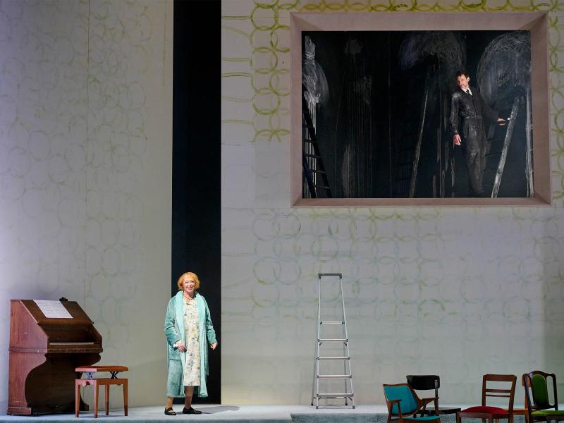 Bühnensituation: Ilse Ritter steht in einem grün-blauen Bademantel auf der Bühne und lächelt. Links von ihr ein Klavier aus Holz. Rechts von ihr eine Leiter und mehrere Stühle. Im Hintergrund eine hohe weiße Wand mit aufgemalten grünen Kreisen. Die Wand hat eine große rechteckige Öffnung. In der Öffnung ist es dunkel, ein Mann in einem schwarzen Anzug auf einer Leiter ist zu sehen, der zu ihr Ilse Ritter schaut. Die schwarze Wand hinter ihm ist mit verwischter Kreide bemalt.