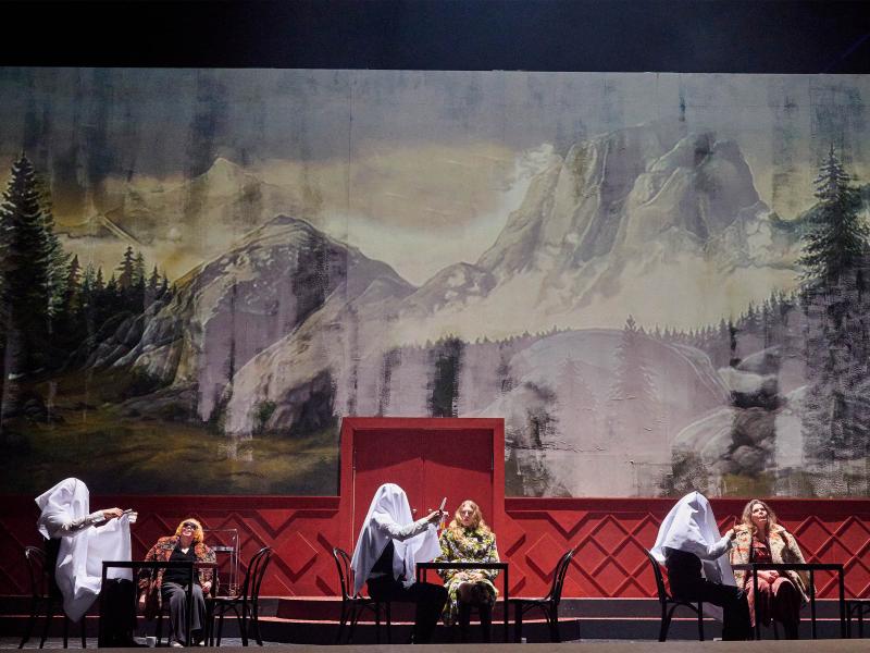 Bühnensituation: Ilse Ritter, Alberta von Polnitz und Julia Wieninger sitzen an drei kleinen Tischen darauf. Mit ihnen am Tisch jeweils ein Mann in weißem Hemd und schwarzer Weste. Die Männer haben weiße Laken über den Kopf gestülpt und halten synchron etwas in ihren Händen. Im Hintergrund ist eine große Wandmalerei von einer Berglandschaft mit Tannen zu sehen.