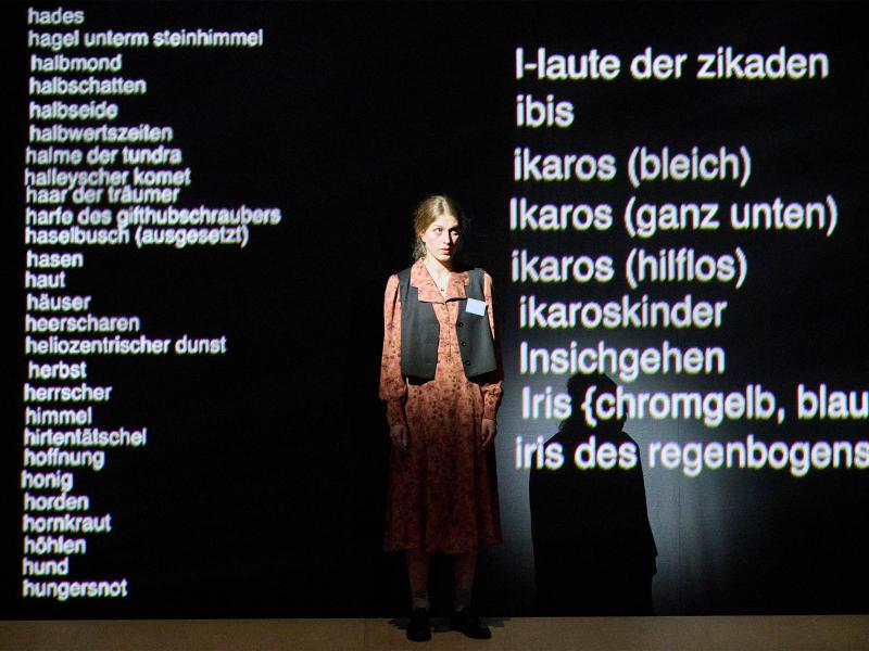 Bühnensituation: Alberta von Polnitz steht in einem gemusterten orangefarbenen Kleid mit schwarzer Weste auf der Bühne und schaut geradeaus. Links und rechts von ihr werden auf eine schwarze Wand untereinander weiße Worte projiziert: halbmond, halbschatten, halbseide, halbwertszeiten ….