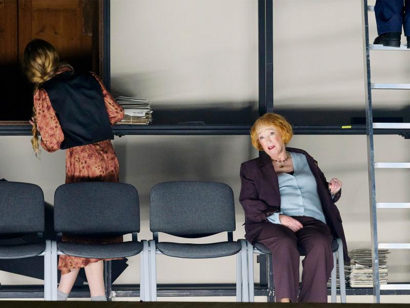 Bühnensituation: Ilse Ritter sitzt auf einem Stuhl. Neben ihr in einer Reihe weitere Stühle. Im Hintergrund steht ein großes Metallgestell, an dem Alberta von Polnitz mit dem Rücken zu ihr steht. Rechts steht eine Leiter, es sind zwei Schuhe und blaue Hosenbeine zu sehen.