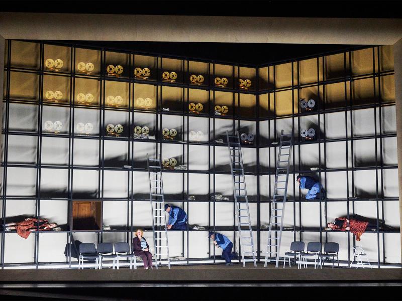 Bühnensituation: Über das Bühnenbild erstreckt sich ein Metallgestell, welches so angeordnet ist, dass sich regalartige Fächer bilden. In den oberen Fächern sind mehrere Tonbandgeräte zu sehen. Etwas weiter unten knien verteilt drei Männer in einzelnen Regalfächern.  Alberta von Polnitz und Julia Wieninger liegen jeweils links und rechts in Fächern, Ilse Ritter sitzt auf einem Stuhl in einer Stuhlreihe.