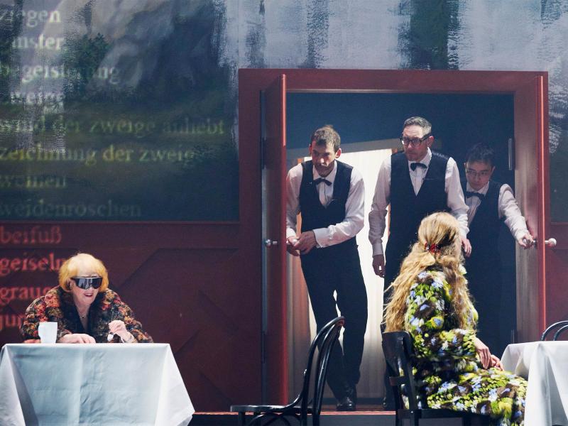 Bühnensituation: Ilse Ritter sitzt an einem Tisch mit einer weißen Tischdecke und einem weißen Becher darauf. Sie trägt eine große Sonnenbrille. Neben ihr Alberta von Polnitz an einem weiteren, identischen Tisch. Im Hintergrund ist eine große Wandmalerei von einer Berglandschaft mit Tannen zu sehen. Inmitten befindet sich eine rote Tür, aus der drei Männer mit schwarzen Hosen, weißen Hemden, schwarzer Weste und Fliege herauskommen, sie schauen zu Ilse Ritter. 