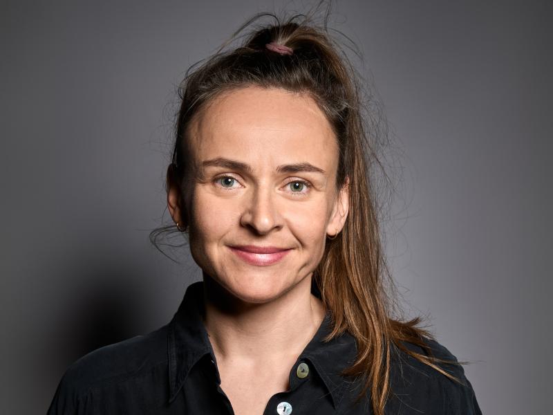 Henni Jörissen steht vor einem neutralen Hintergrund. Sie trägt eine schwarze Bluse, lange, zu einem Pferdeschwanz hochgebundene mittelbraune Haare und schaut lächelnd in die Kamera.