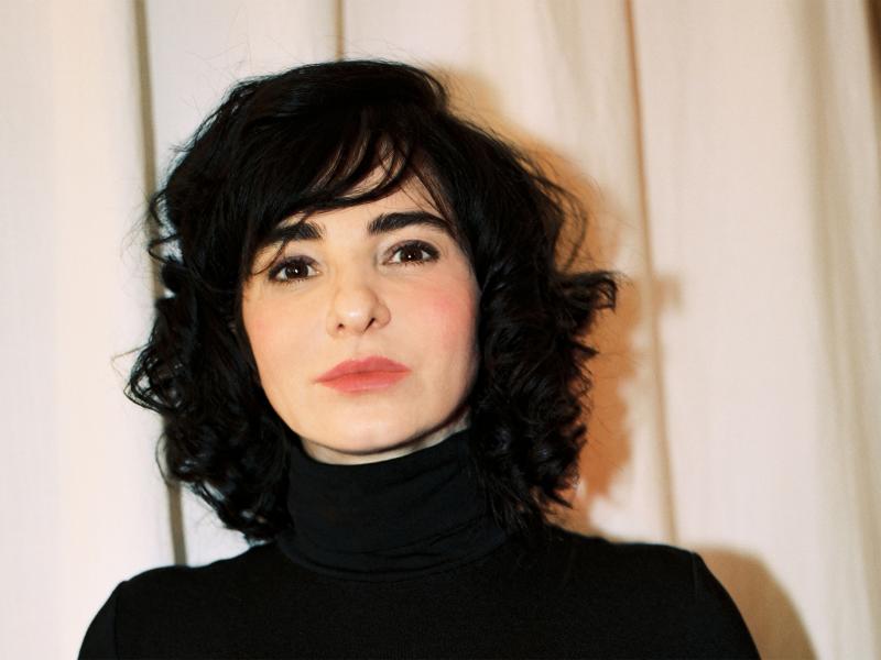 Mely Kiyak steht vor einem hellen Hintergrund in einem Innenraum. Sie trägt ein schwarzes hochgeschlossenes Oberteil, schulterlange schwarze Haare und schaut in die Kamera.